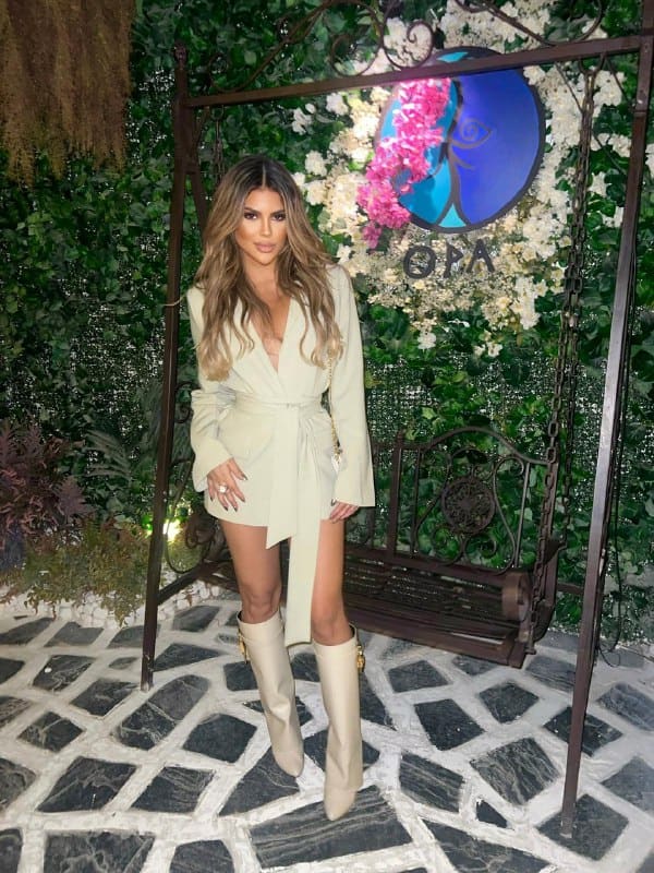 A woman in a cream belted trench dress and knee-high boots stands before a floral wall and metal swing on tiled flooring.