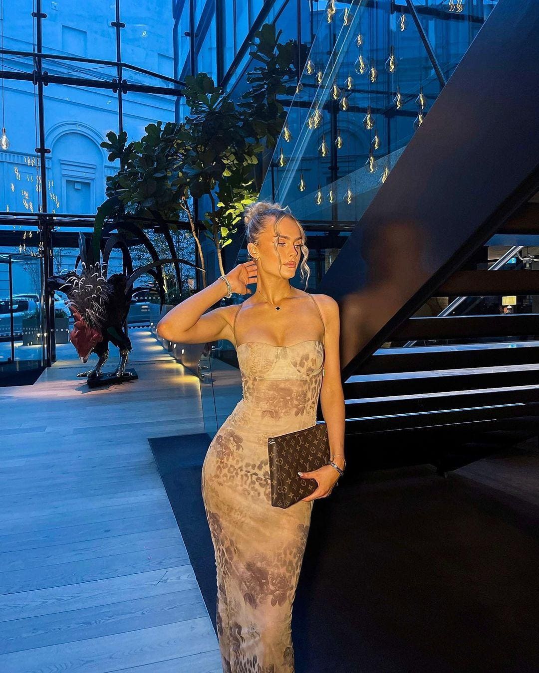 A woman in a sheer floral strapless gown stands in a glass lobby holding a clutch, with a bird sculpture behind her.