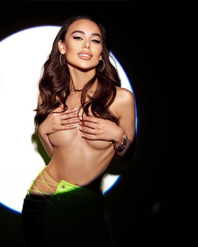A woman covers her chest with both hands, wearing a black skirt with green lacing, in front of a circular light.
