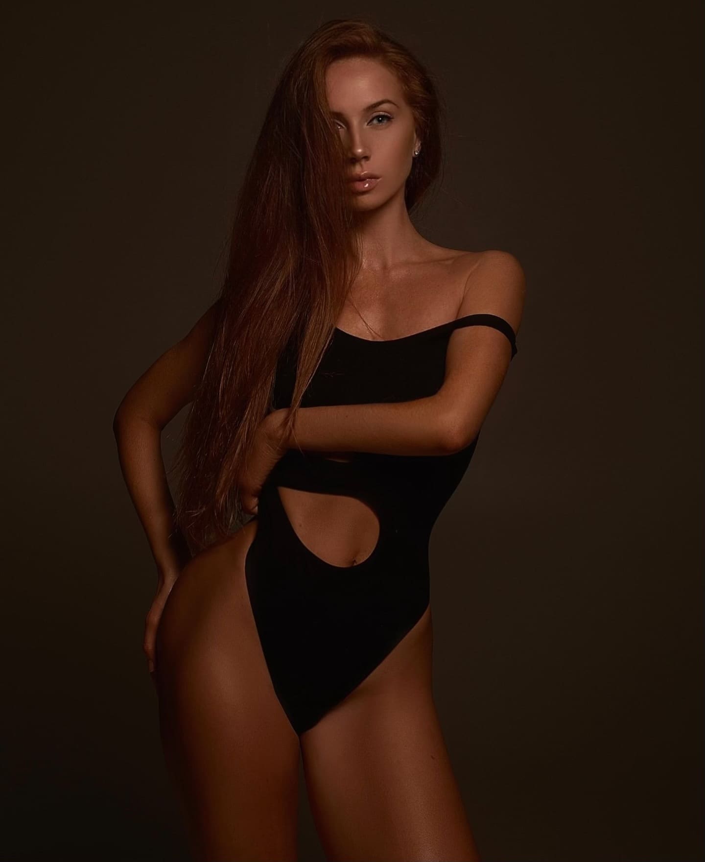 A woman stands in a studio wearing a black one-shoulder bodysuit with a midriff cutout, her arms crossed in front of her torso.