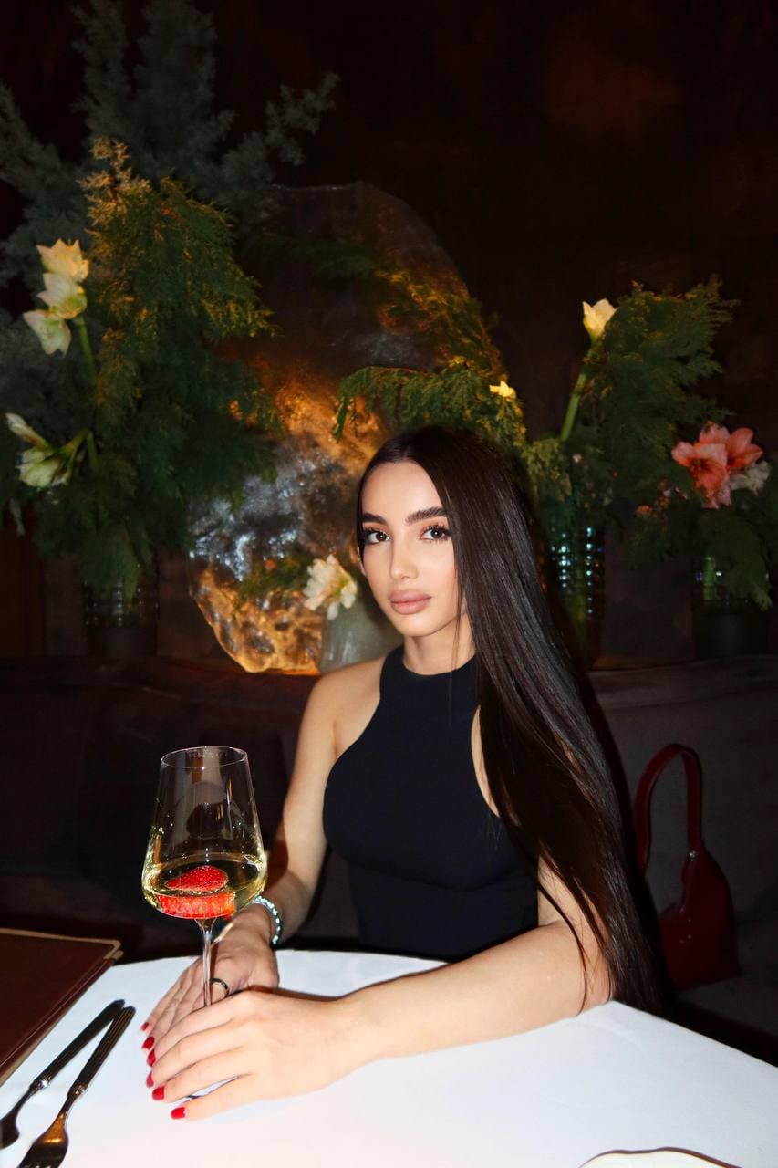 A woman sits at a white-clothed table in a dimly lit room, holding a glass of white wine with a strawberry, wearing a black halter top.