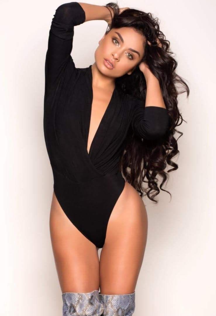 Lana stands against a white background in a black deep-V bodysuit and patterned knee-high boots, hands raised to her long dark wavy hair.