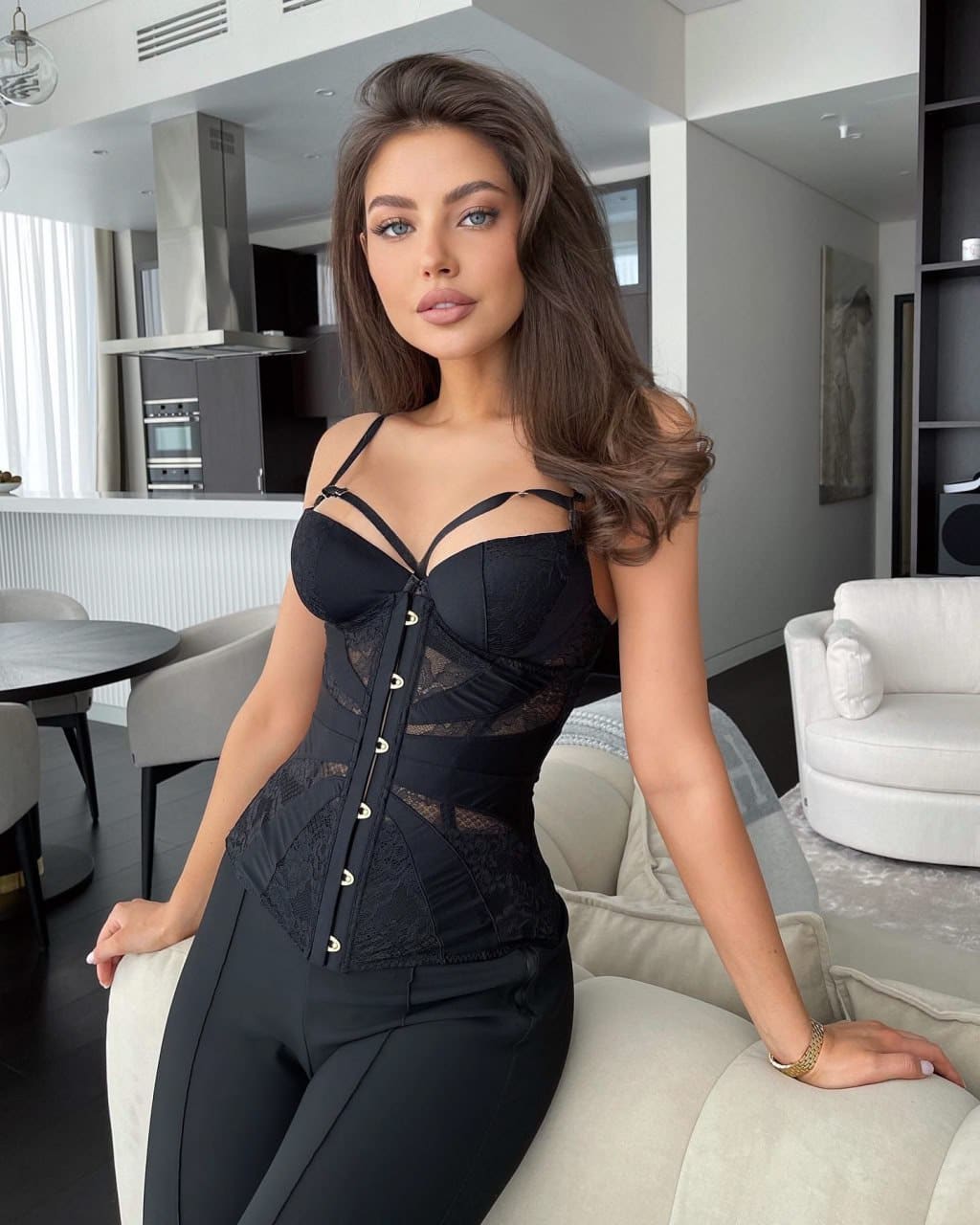 A woman sits on a light sofa in a modern living room, wearing a black lace corset top with front lacing and black trousers.