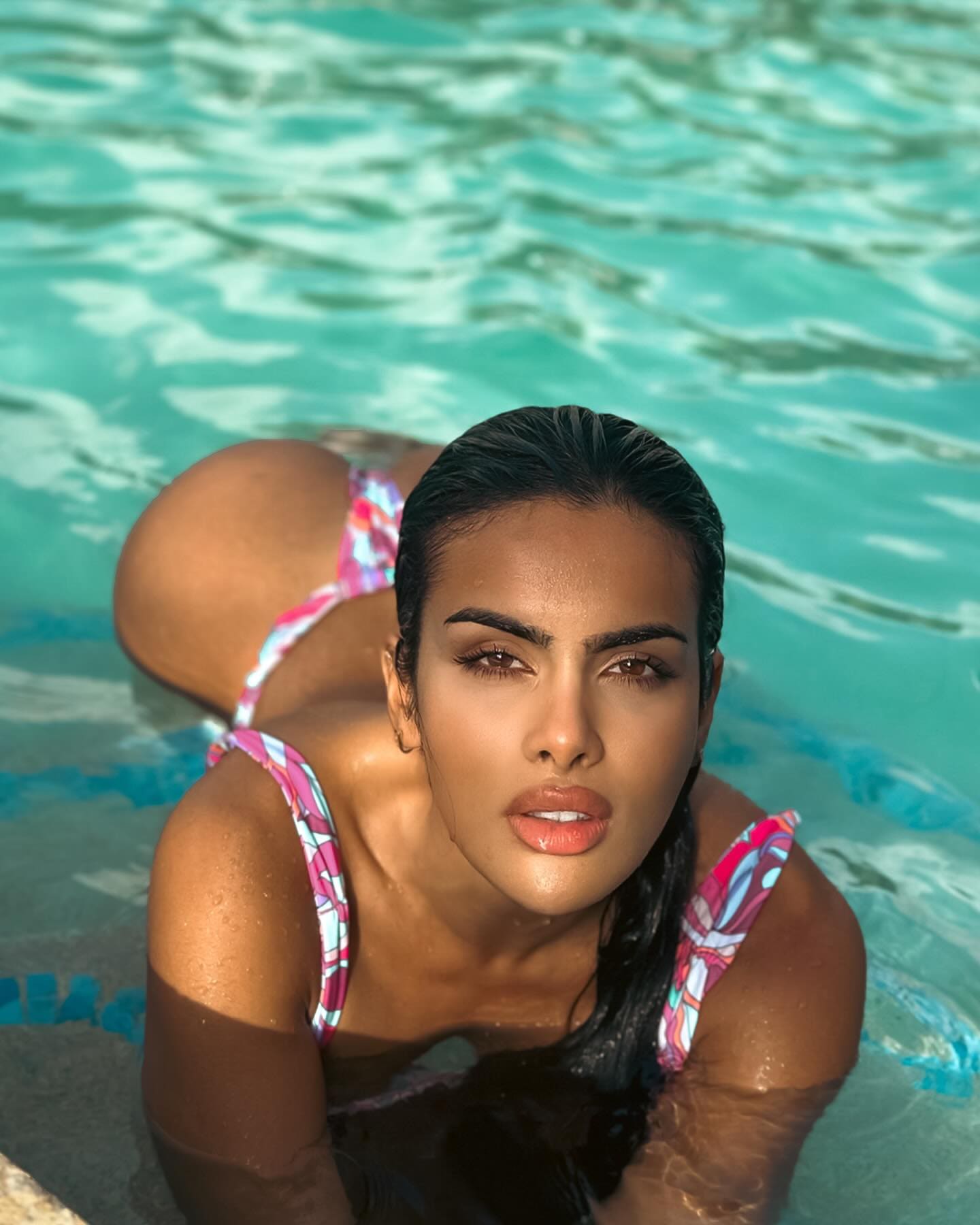 A woman lies on her stomach in a turquoise pool in a pink and white patterned bikini, upper body above water, legs bent behind her.