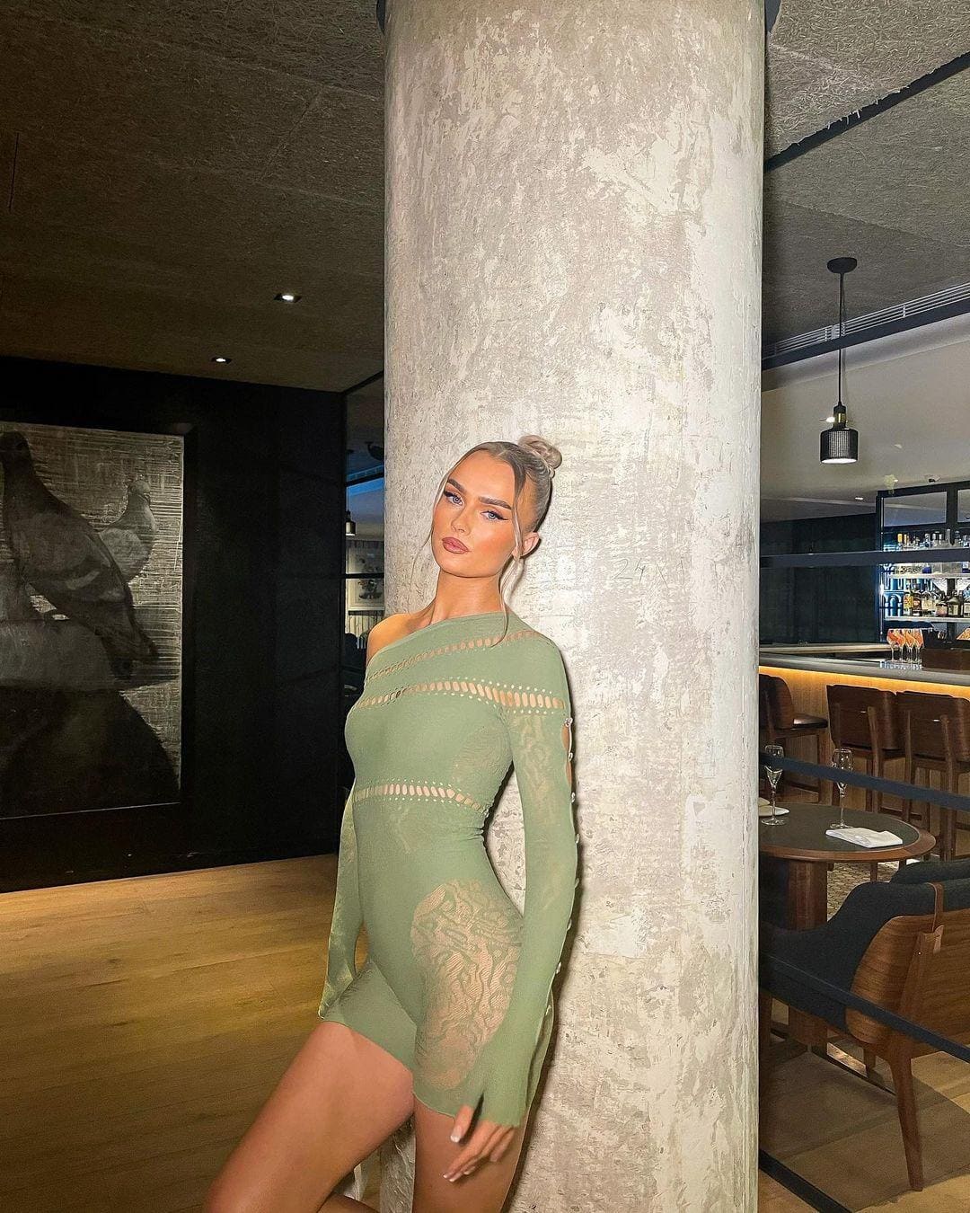 A woman in a sage green bodysuit with lace panels leans against a concrete pillar in a dimly lit restaurant.