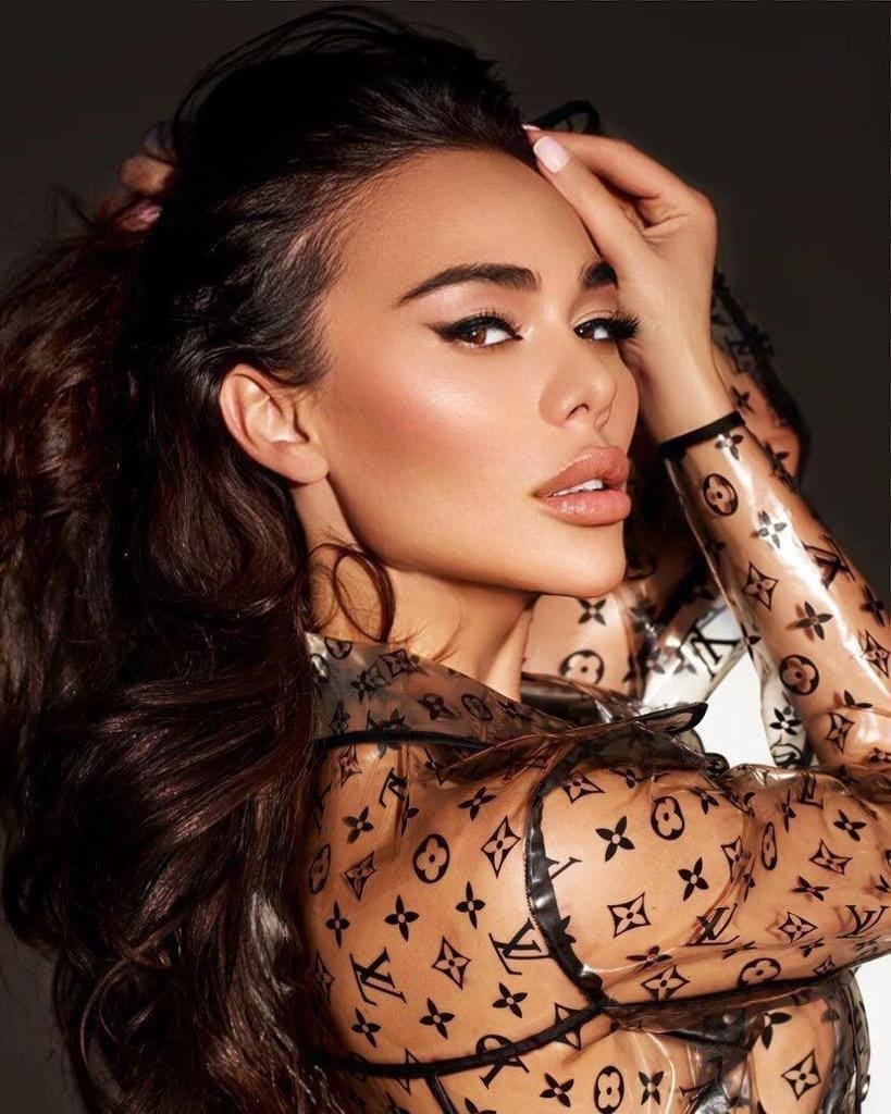 Oleksandra with long dark wavy hair wears a sheer monogram top, hand raised to her head, against a dark background.