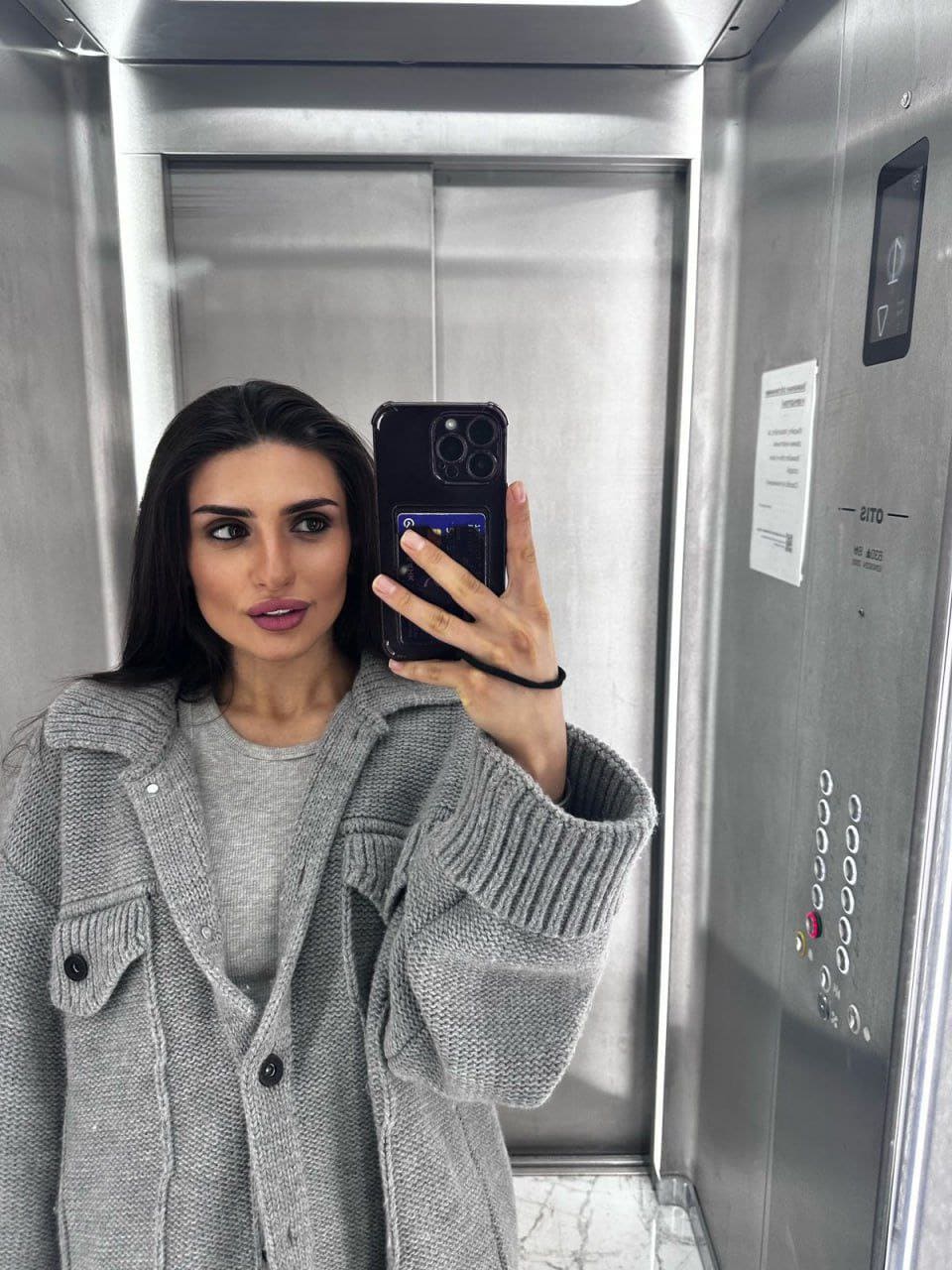 Mirror selfie in an elevator showing a woman in a grey knit cardigan over a grey t-shirt holding a phone.