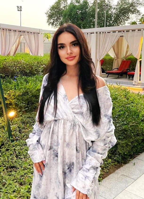 A woman stands in a garden wearing a loose, off-the-shoulder grey and white marbled dress, positioned in front of green hedges and a pergola.