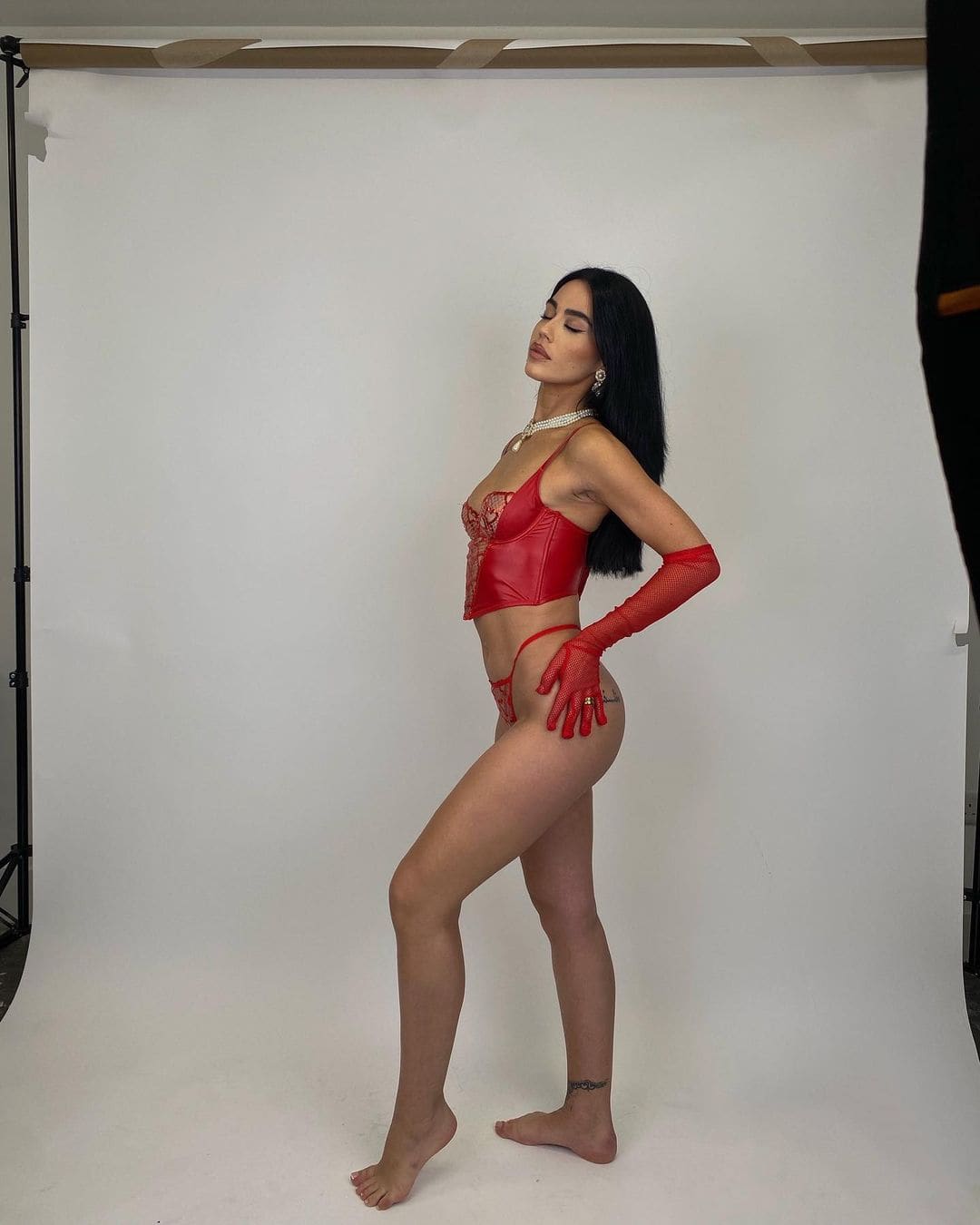Barefoot in a studio, a woman tilts her head back wearing a red lace-and-leather bra top, red underwear, and long red mesh gloves.