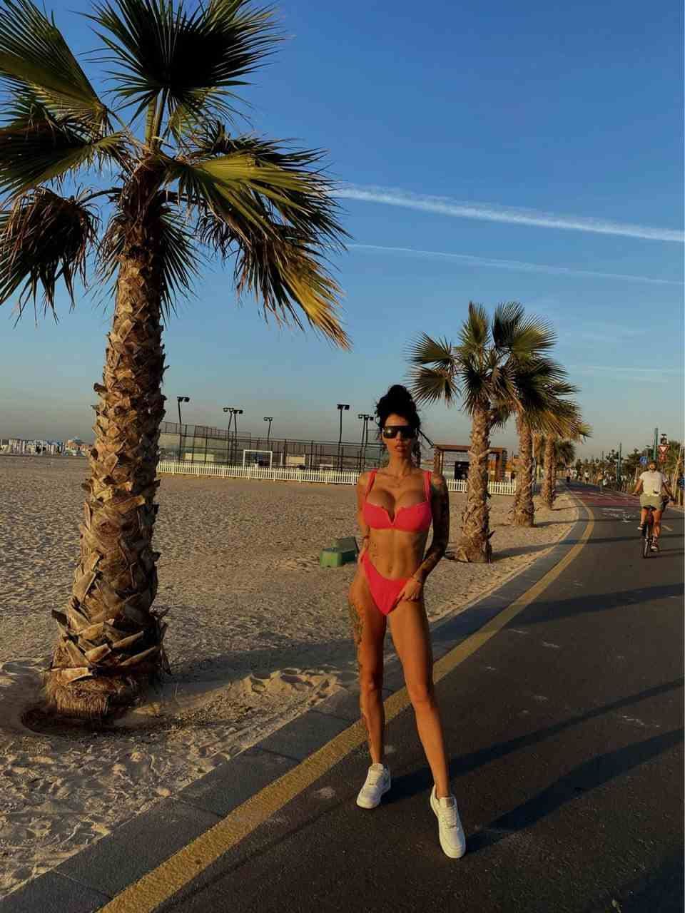 A woman in a pink bikini and white sneakers stands on a path beside a sandy beach, with a cyclist in the background.