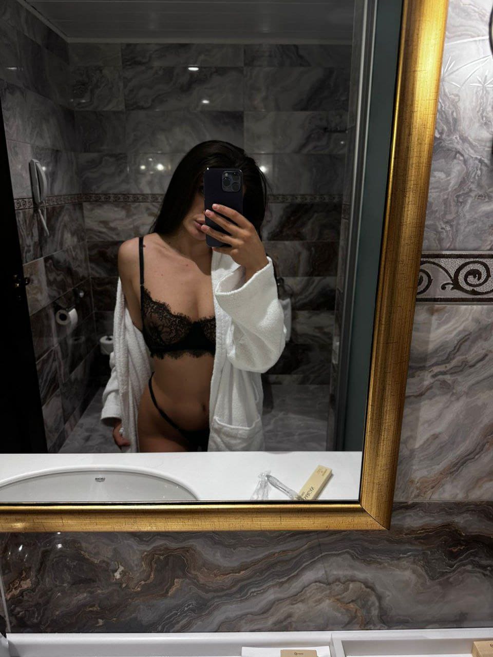 Mirror selfie in a marble bathroom with an open white bathrobe revealing black lace lingerie underneath.