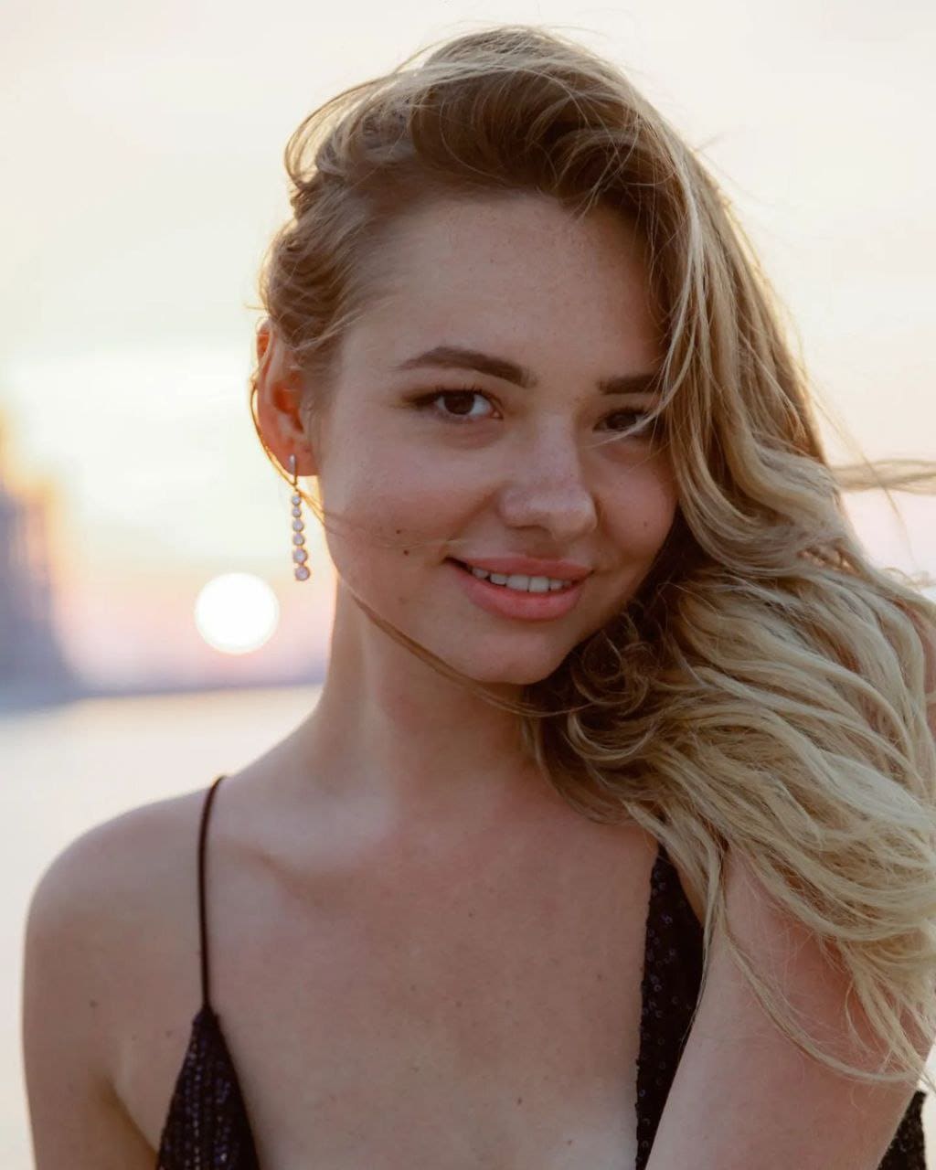 Katusha stands outdoors in a black sequined top with thin straps and pearl earrings, with a blurred sunset and water behind her.