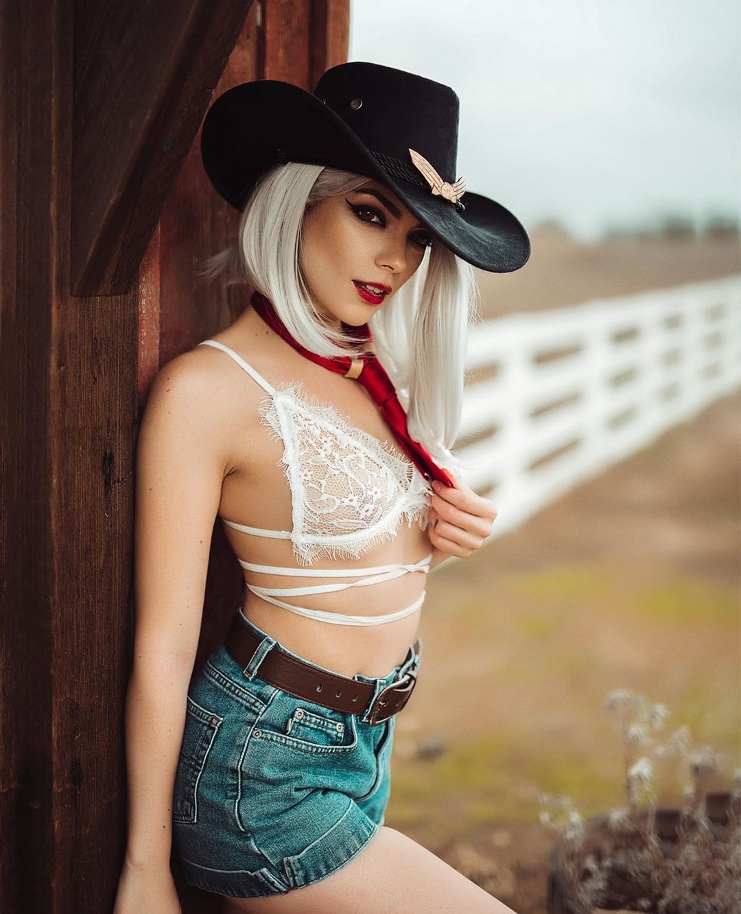 Person in a cowboy hat, white lace bralette, red scarf, and denim shorts leans against a wooden post in a field with a white fence in the background.