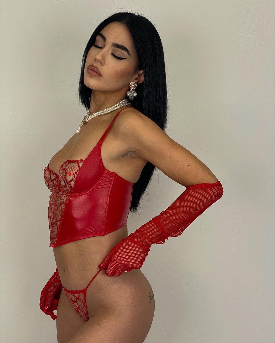 A woman in profile wears a red lace and patent-leather bustier, matching briefs, red mesh gloves, and pearl jewelry against a light wall.