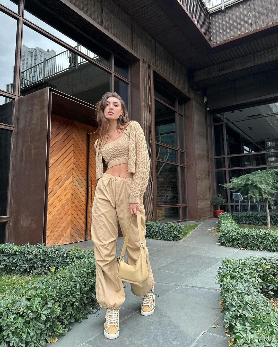 A woman stands on a paved walkway before a modern building, wearing a beige knit crop top, cardigan, tan trousers, and holding a shoulder bag.