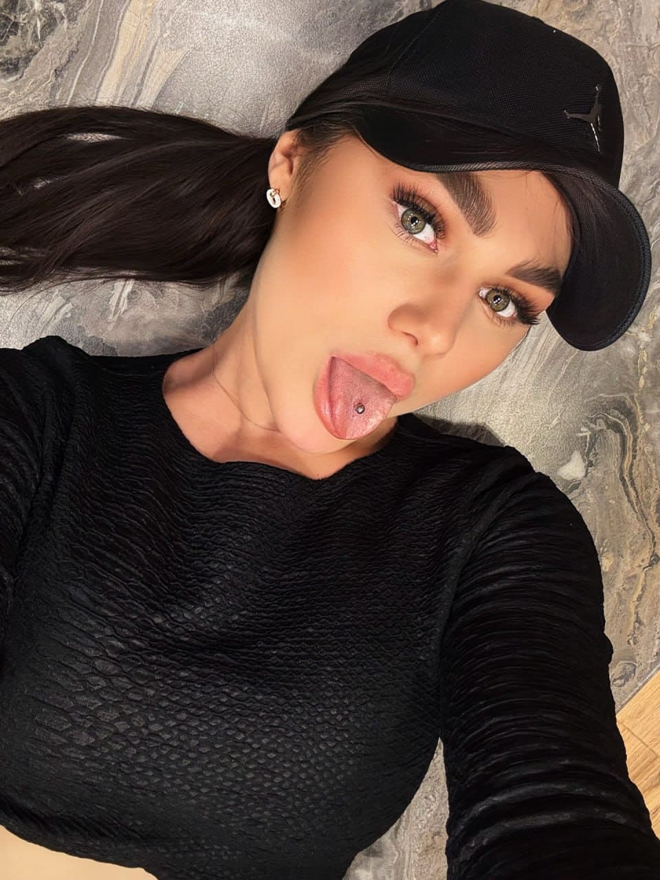Lying on a gray marbled surface in a black baseball cap and textured top, sticking her tongue out toward the camera.