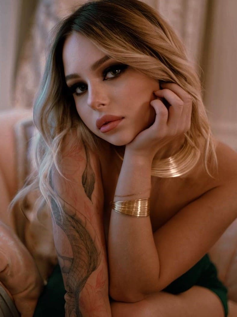 A blonde woman with a floral arm tattoo sits on a tufted sofa in a dark green garment, resting her chin on her hand adorned with bangles.