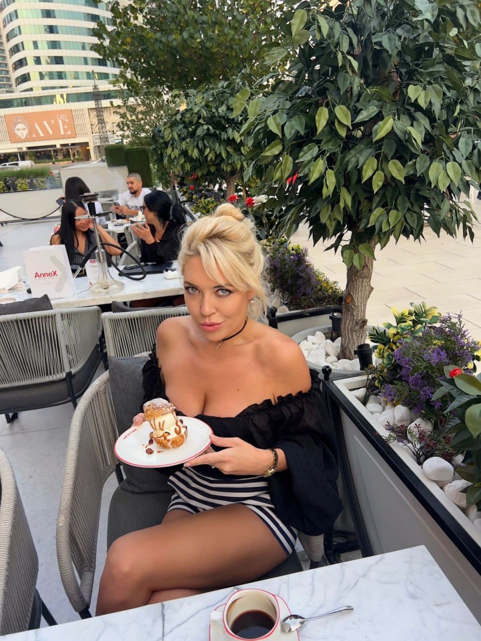 Stasya sits at an outdoor cafe table in a black off-shoulder top and striped skirt, holding a plate of pastry with coffee nearby.