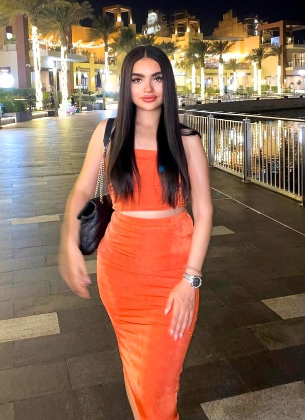A woman stands on a paved walkway at night in an orange tube top and long skirt, carrying a black bag, with illuminated buildings and palm trees behind.