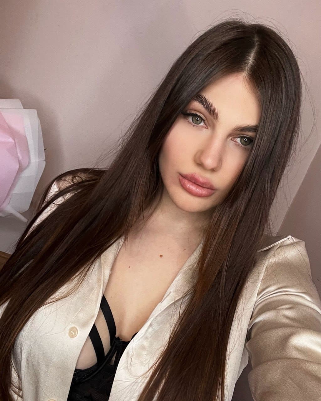 Close-up selfie in an open cream satin blazer over a black strappy top, against a plain light wall.