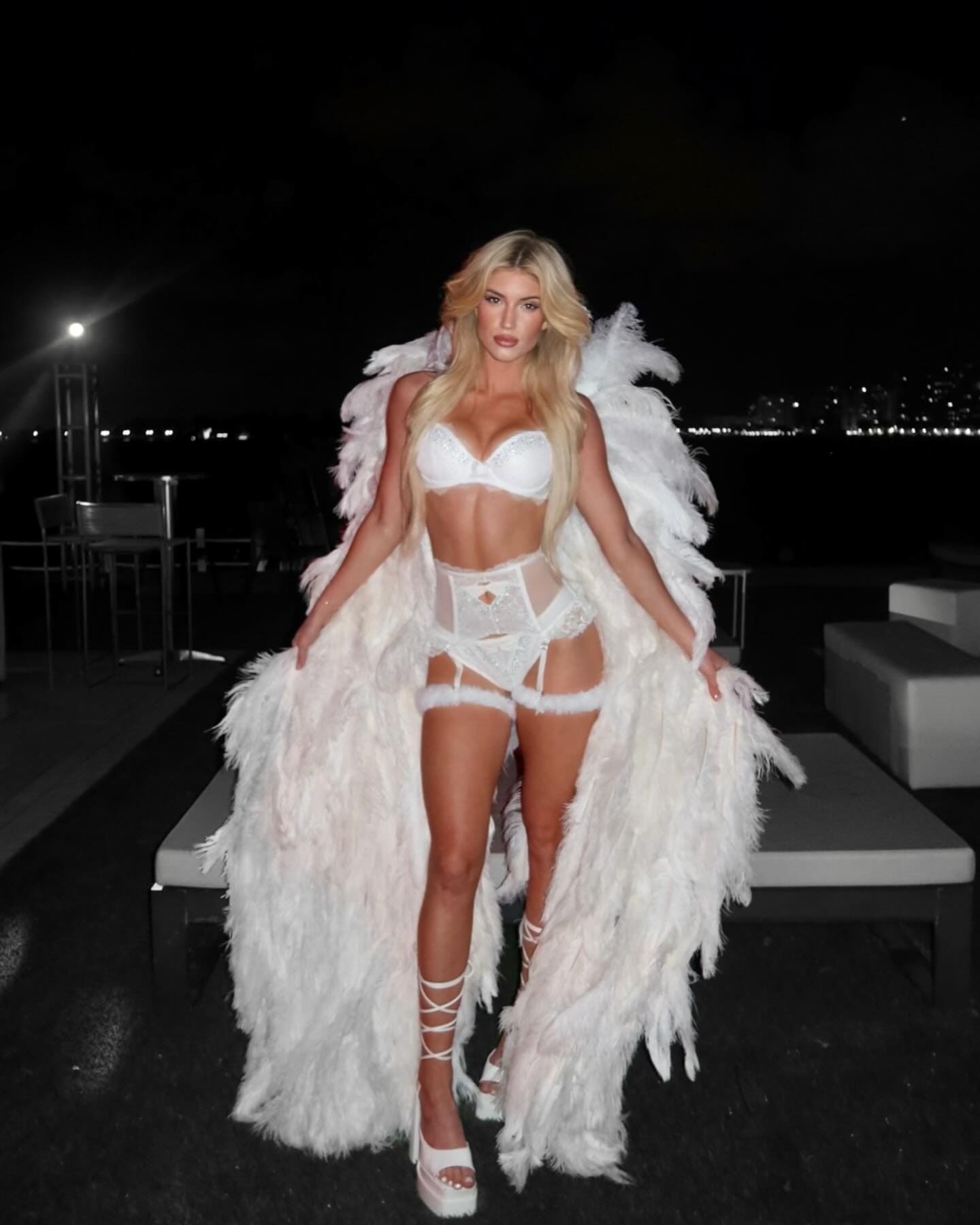 A blonde woman stands outdoors at night in a white bra, sheer lace bodysuit, and feathered white coat, paired with strappy white platform heels near lounge furniture.