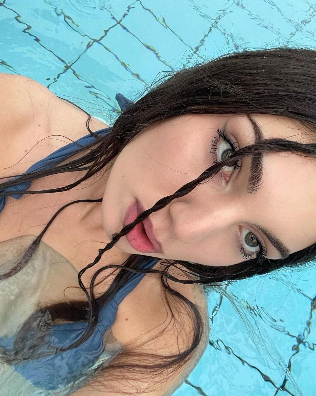 Close-up selfie of a woman with wet hair floating in a blue-tiled pool, wearing a blue one-piece swimsuit with a sheer chest panel.
