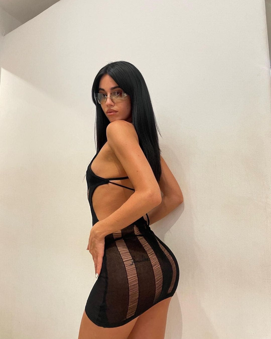 In profile against a white wall, a woman with tinted glasses wears a black cut-out top and a sheer striped mini skirt.