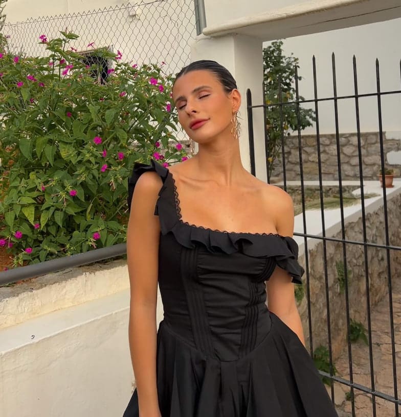 Standing outdoors by a white wall and flowering bush, she wears a black one-shoulder ruffled dress with eyes closed and head tilted.