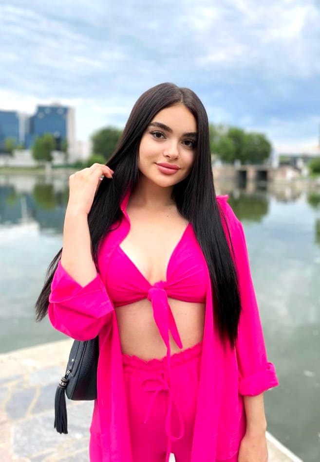 Vika stands outdoors by water, wearing a pink open shirt over a tie-front top and high-waisted pants, with one hand raised to her hair.