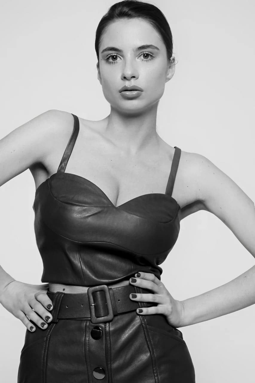 A woman stands with hands on hips in a dark leather bustier and matching skirt with a wide belt, against a light background.