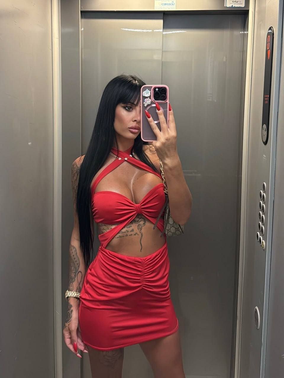 A woman with tattoos takes a mirror selfie in an elevator, wearing a red strappy halter top and mini skirt, holding a pink phone.