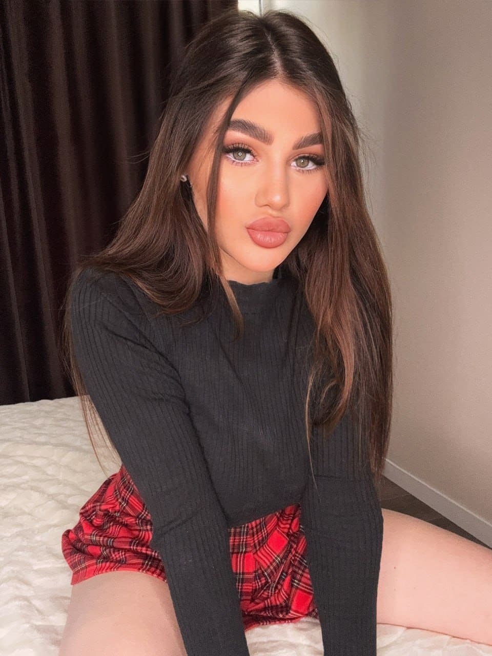 Emilia sits on a white bed in a black ribbed top and red plaid skirt, with a dark curtain behind her.