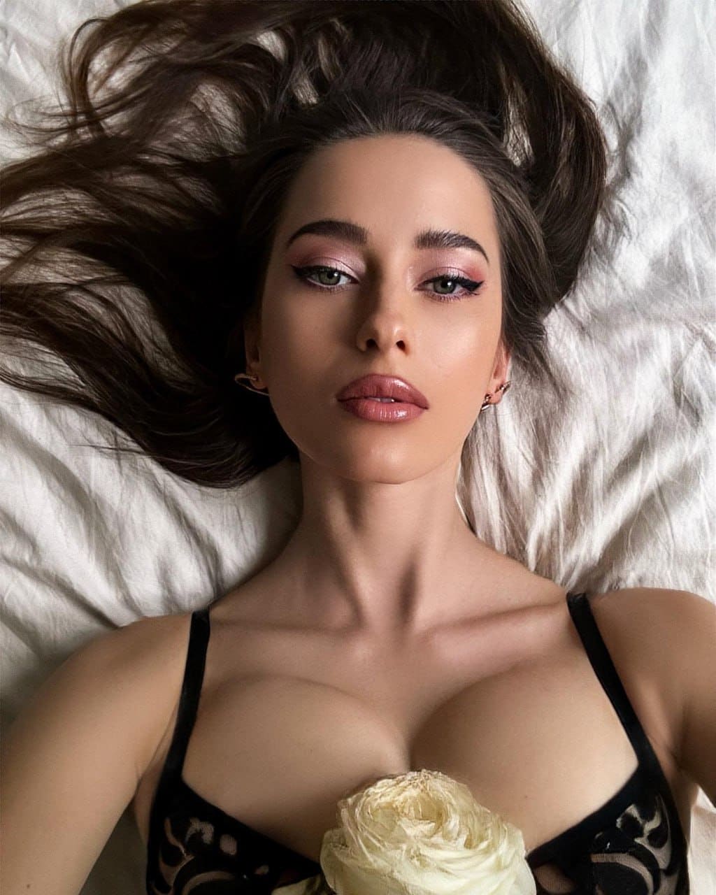 A woman lies on white bedding in a black lace bra, holding a pale rose to her chest while looking up at the camera.