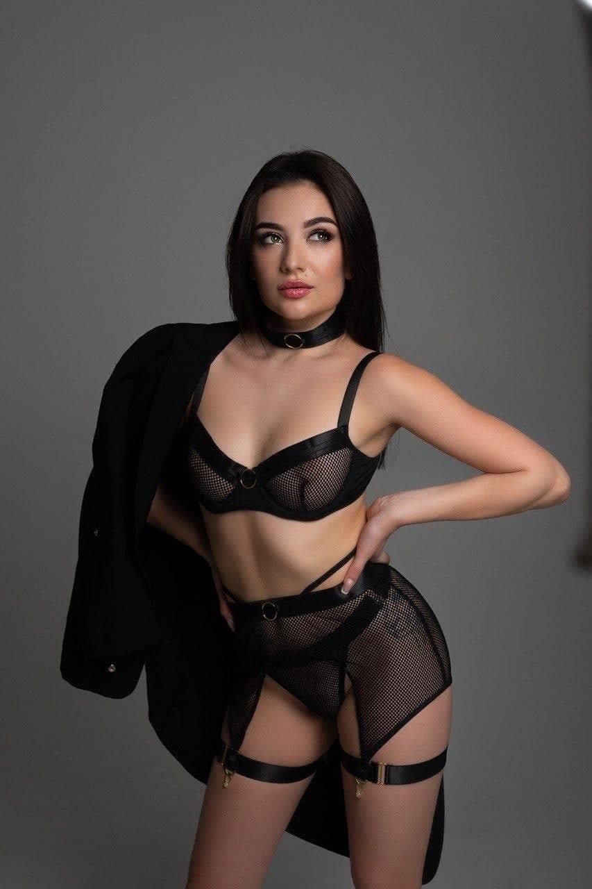 Brienne stands against a gray backdrop in black mesh lingerie and a blazer draped over one shoulder, hand on hip.
