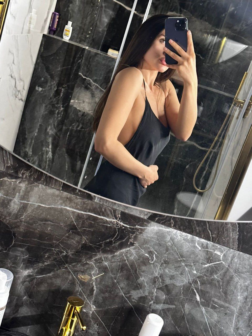 Mirror selfie in a dark marble bathroom wearing a black spaghetti-strap slip dress, phone covering face.