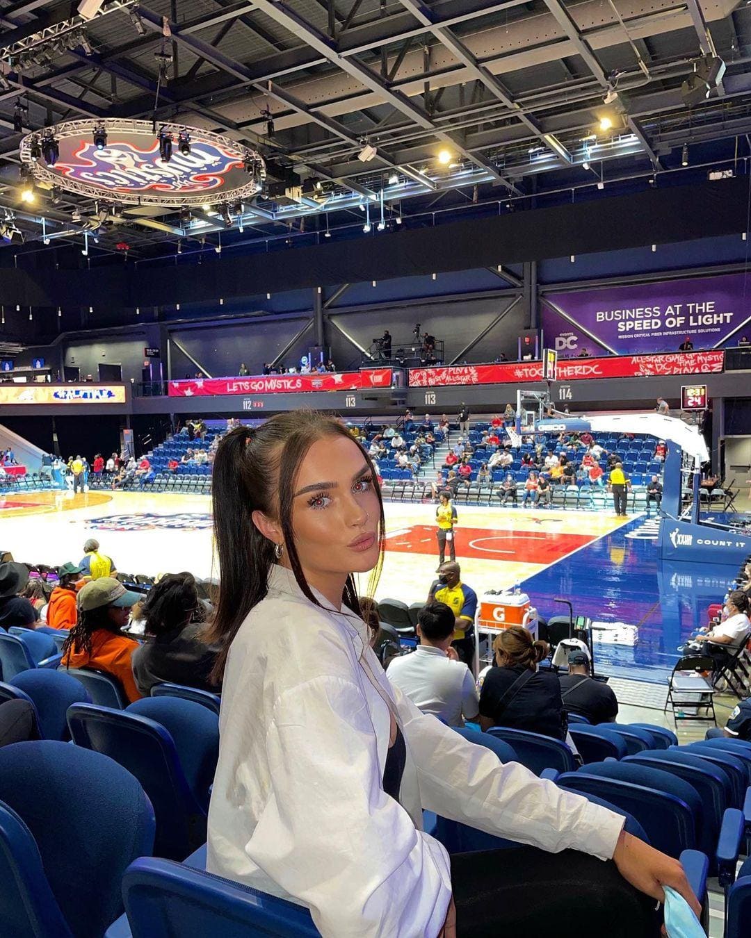 Sasha sits in blue stadium seats wearing an open white shirt over a dark top, looking back over her shoulder.
