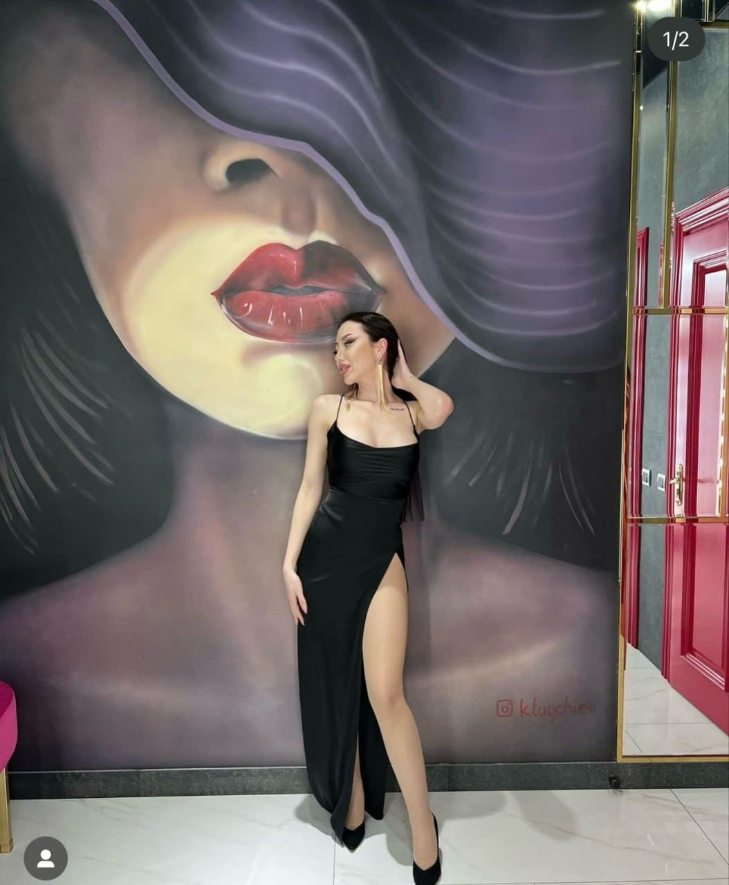 A woman in a black spaghetti-strap gown with a thigh slit stands before a face mural, touching her hair.