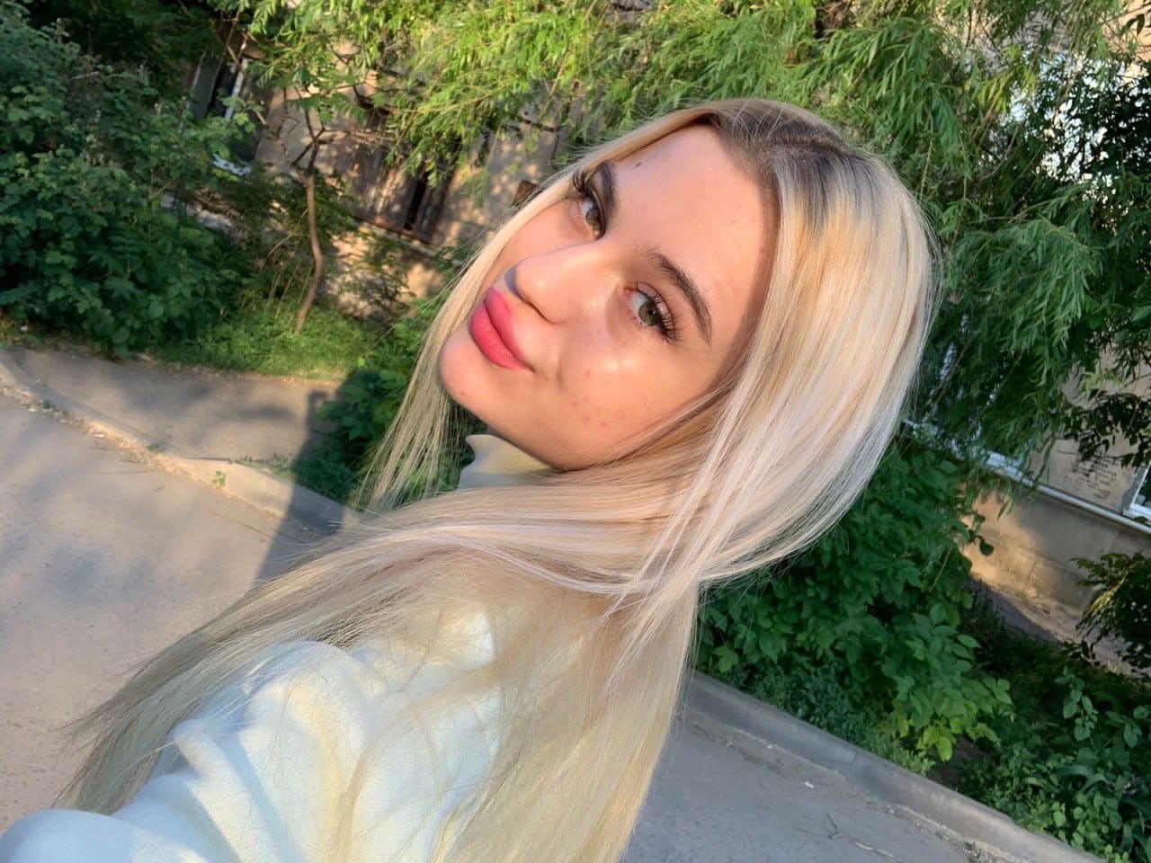 Monica with long blonde hair and pink lipstick tilts her head outdoors on a paved path near green foliage and a building.