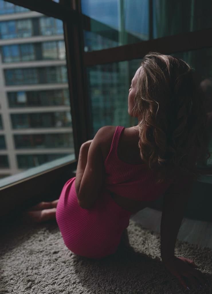 A woman in a pink ribbed two-piece outfit sits on a shaggy rug, looking out a window at a city building.