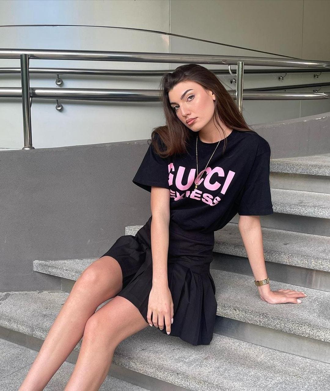 A woman sits on concrete steps near a metal railing, dressed in a black t-shirt with pink lettering and a black skirt.