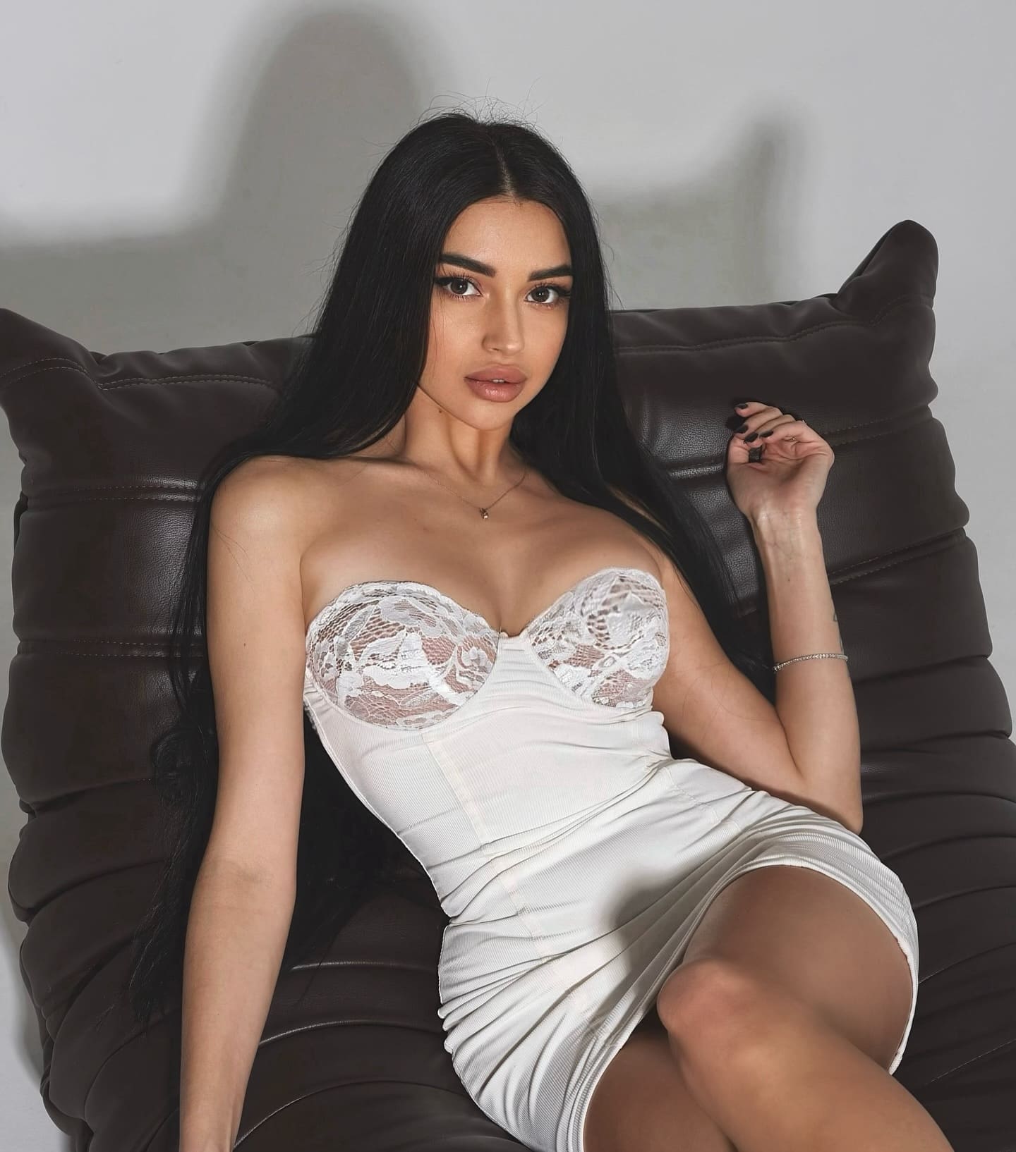A woman reclines on a dark leather chair wearing a short white strapless dress with lace detailing, hand raised near her shoulder.