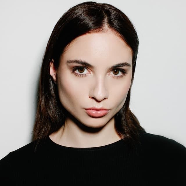 Kaya faces the camera in a close-up, wearing a black crew-neck top against a plain light gray background.