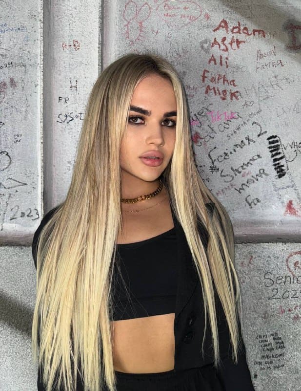 A blonde woman in a black cropped top and buttoned jacket with a gold chain stands before a concrete wall covered in handwritten names.