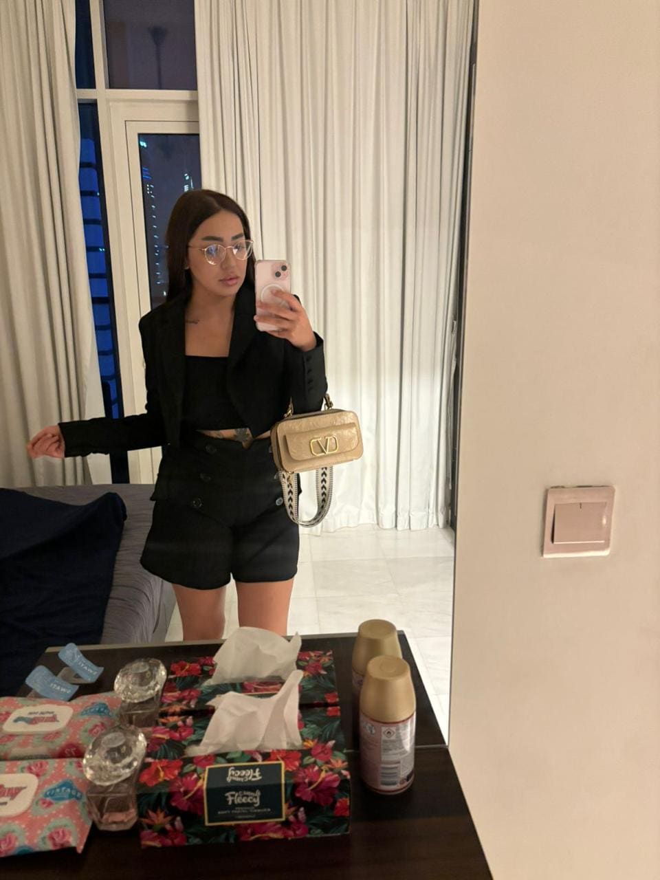 Woman with glasses in a black blazer, crop top, and shorts with a crossbody bag, taking a mirror selfie in a room with white curtains.