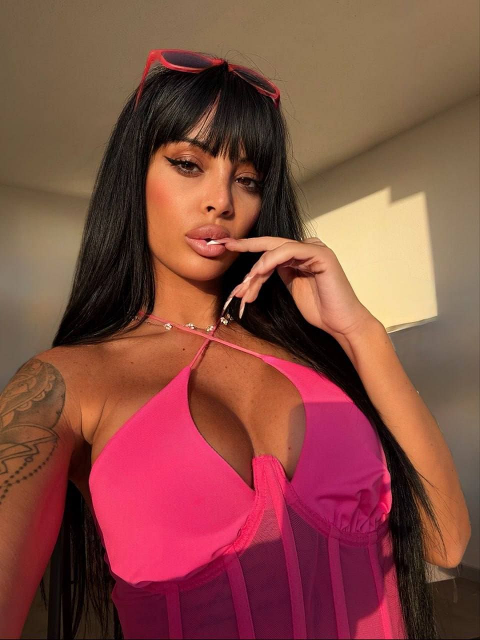 Emily wears a bright pink halter top with a sheer panel, posing indoors with one hand near her mouth and sunglasses on her head.