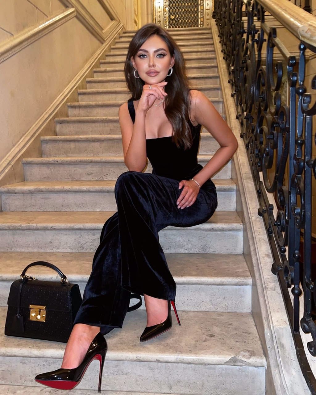 A woman sits on a marble staircase in a black sleeveless top, velvet wide-leg trousers, and patent heels, with a handbag beside her.