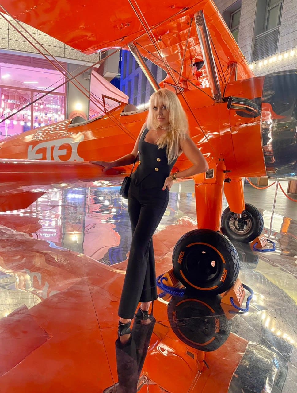 A blonde woman in a black jumpsuit with gold buttons stands beside a bright orange biplane on a reflective floor, one hand on her hip.