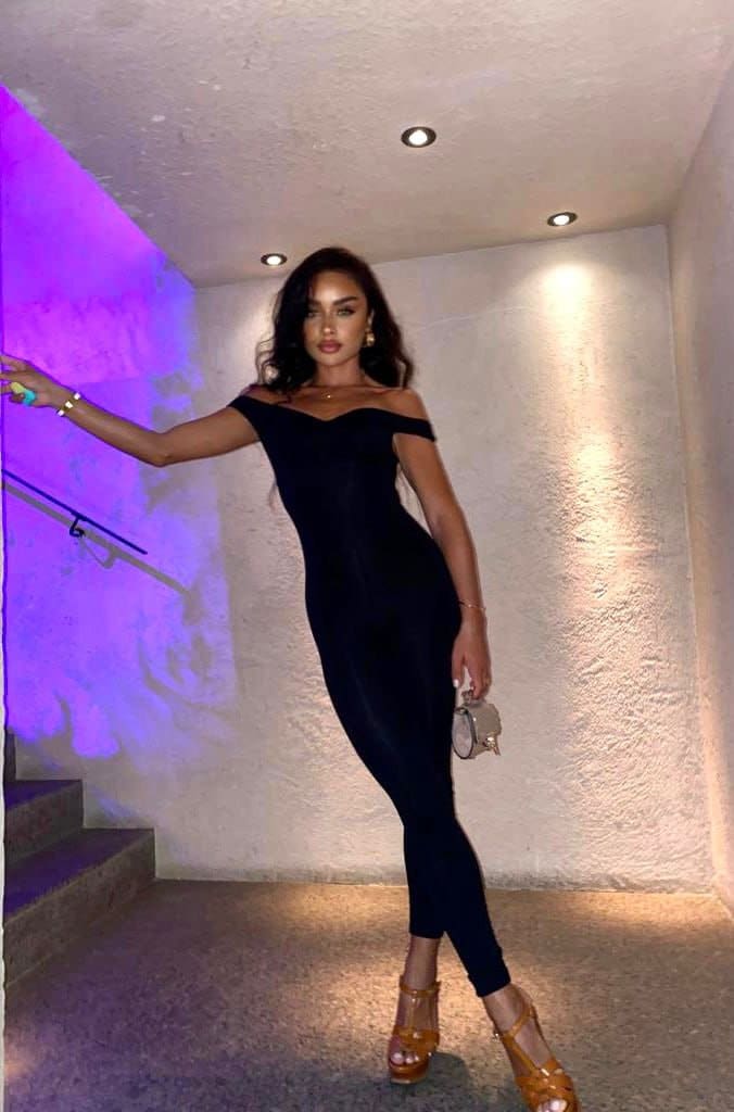 A woman in a black off-shoulder dress and tan platforms stands in a purple-lit stairwell, holding a metallic clutch and resting a hand on a railing.