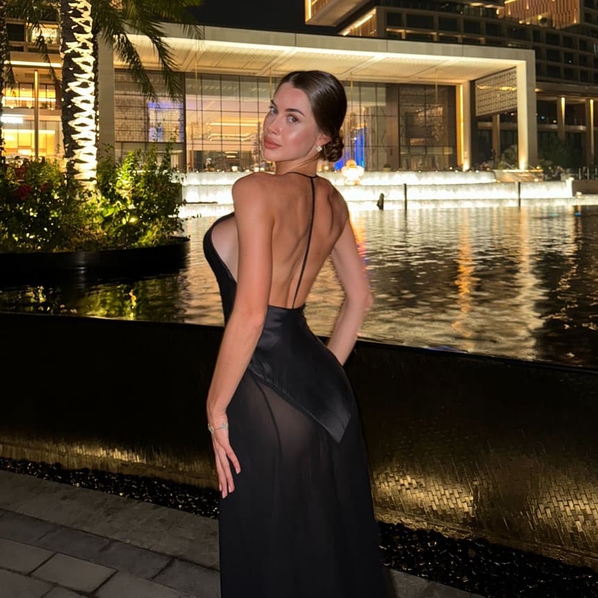 Profile view of a woman in a black gown with an open back, standing beside a lit water feature at night.