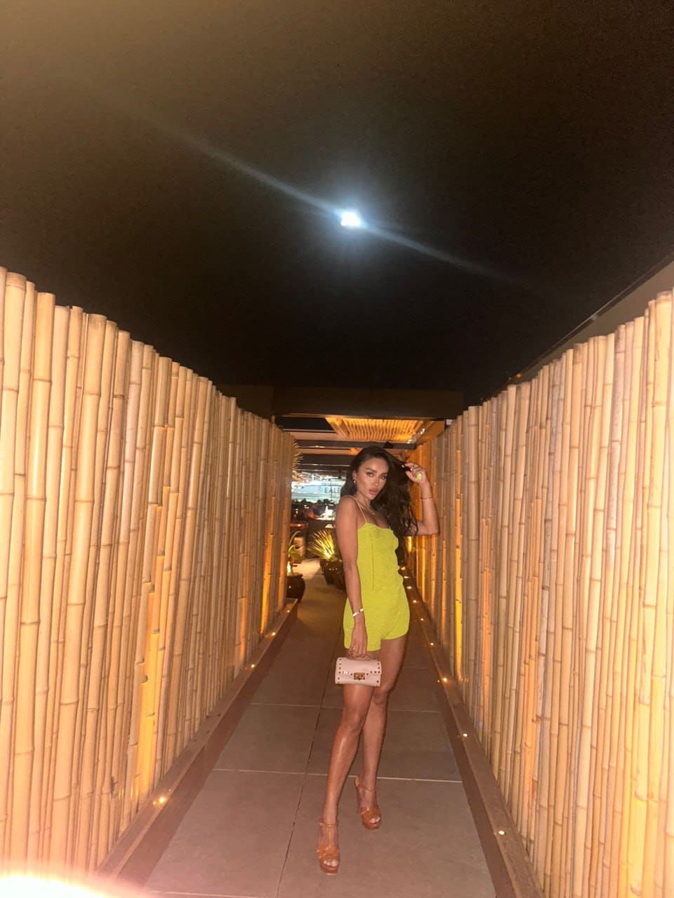 A woman in a lime-green sleeveless dress and tan heels stands in a bamboo-lined corridor, holding a light handbag and touching her hair.